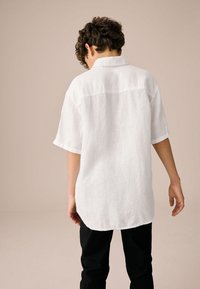 Weißes Leinenhemd mit kurzen Ärmeln und Kragen, mit geradem Saum und feiner Struktur. Kombiniert mit engen schwarzen Hosen.