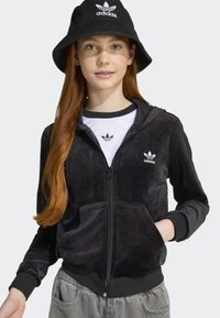 Black zip-up velour jacket with hood, ribbed cuffs, and front pockets. White Adidas logo at chest and on bucket hat. Gray pants visible below.