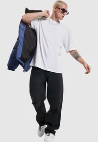 Chaqueta acolchada negra con forro azul, camiseta blanca y jeans negros desgastados. El modelo lleva gafas de sol y zapatillas blancas.