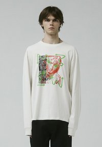 Camisa de manga larga color crema con un estampado gráfico en negro, rojo y verde, con elementos abstractos y acentos de texto. Tejido de algodón suave.
