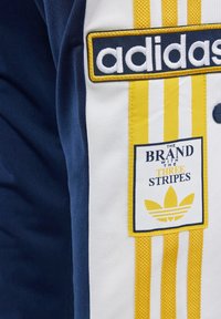 Námořnická bunda s žlutými a bílými pruhy. Vyšívané logo "adidas" a značková nášivka se třemi pruhy. Materiál má hladkou texturu.