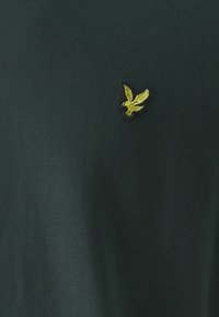 Tmavozelené tričko vyrobené z bavlny, s vyšitým žltým logom orla na ľavej strane hrudníka. Hladká textúra, s krátkymi rukávmi.