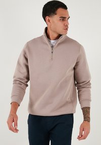 Sudadera beige con cuello de media cremallera, mangas largas, puños acanalados y una textura suave. Llevada sobre una camiseta blanca y combinada con pantalones oscuros.