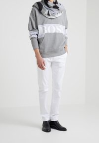 Persona con sudadera con capucha gris con texto "BOSS", bufanda en blanco y negro, pantalones blancos y botines negros de tobillo, de pie en una habitación blanca.