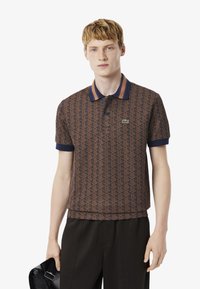 Polo shirt à motifs marron avec un col et des poignets côtes bleu marine, arborant un logo vert et une patte de boutonnage. Tissu léger.