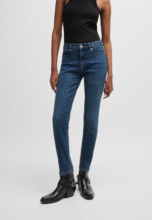 Jeans blu scuro skinny con vita alta, design a cinque tasche e cuciture a contrasto. Indossati con stivali neri alla caviglia e un top nero senza maniche.