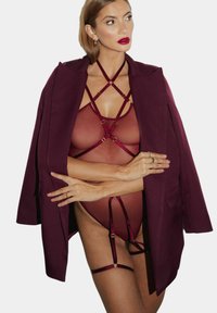 Bordowy płaszcz na przezroczystym body z krzyżującymi się paskami i złotymi akcentami. Przezroczysty materiał o gładkiej fakturze, odsłaniający skórę pod spodem.