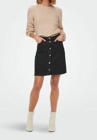 Donna che indossa un maglione di lana beige, una gonna di jeans nera con bottoni davanti e stivali alla caviglia crema, in piedi con le mani nelle tasche.
