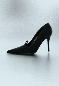 Chaussure à talon haut noire à bout pointu avec talon stiletto sur un fond de surface lisse gris clair.