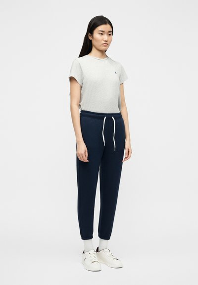 Νεαρή γυναίκα που φορά σκούρο μπλε αθλητικό παντελόνι, γκρι κοντομάνικη μπλούζα, λευκές κάλτσες και λευκά αθλητικά παπούτσια μπροστά σε απλό φόντο.