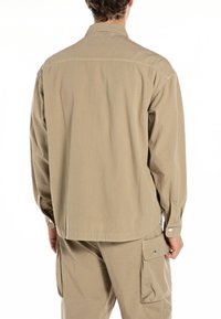 Beige long-sleeve shirt with a back yoke, buttoned cuffs, and a relaxed fit, made from lightweight fabric with a textured finish.