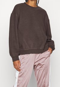 Brązowy polarowy sweatshirt o luźnym kroju, z prążkowanymi mankietami i dołem, haftowanym logo na piersi, w zestawie z jasnoróżowymi spodniami dresowymi.