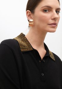 Schwarzer Pulli mit goldbesetztem Kragen, strukturiertem Stoff und Knopfleiste. Die Model trägt große metallische Ohrringe und zeigt das Seitenprofil.