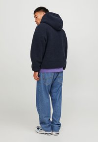 Navy blue fleece hoodie with a textured, fuzzy exterior and a spacious hood, worn over a purple shirt and paired with loose-fitting blue jeans.