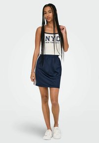 Gonna sportiva blu navy con strisce bianche ai lati, abbinata a una canotta crema con il testo blu "NYC". Il tessuto sembra liscio e leggero.