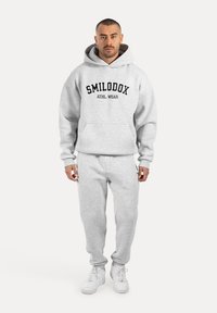 Szara bluza z kapturem i dresowe spodnie z kieszenią z przodu, z czarnym logo "SMILODOX ATHL. WEAR", w komplecie z białymi sneakersami.