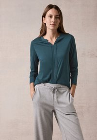 Türkises Langarmshirt mit Knopfblende und grauen Jogginghosen mit Drawstring-Taille und Taschen, vor einem neutralen Hintergrund.