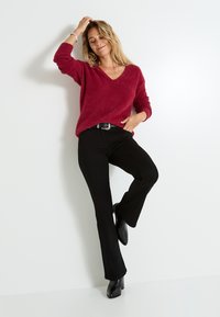 Pull en V rouge texturé associé à un pantalon noir évasé et des bottes noires à talons. L'ensemble est agrémenté d'une ceinture à boucle argentée.