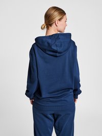 Vrouw met blond haar in een lage knot, gekleed in een navy-blauwe hoodie en bijpassende broek, met de rug naar de camera tegen een effen lichte achtergrond.