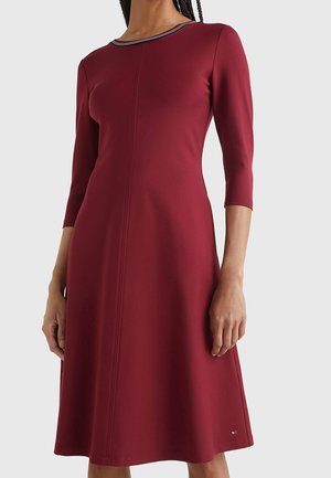 Kobieta w bordowej sukience do kolan z długim rękawem, dopasowaną talią oraz paskowanym detalem przy dekolcie.