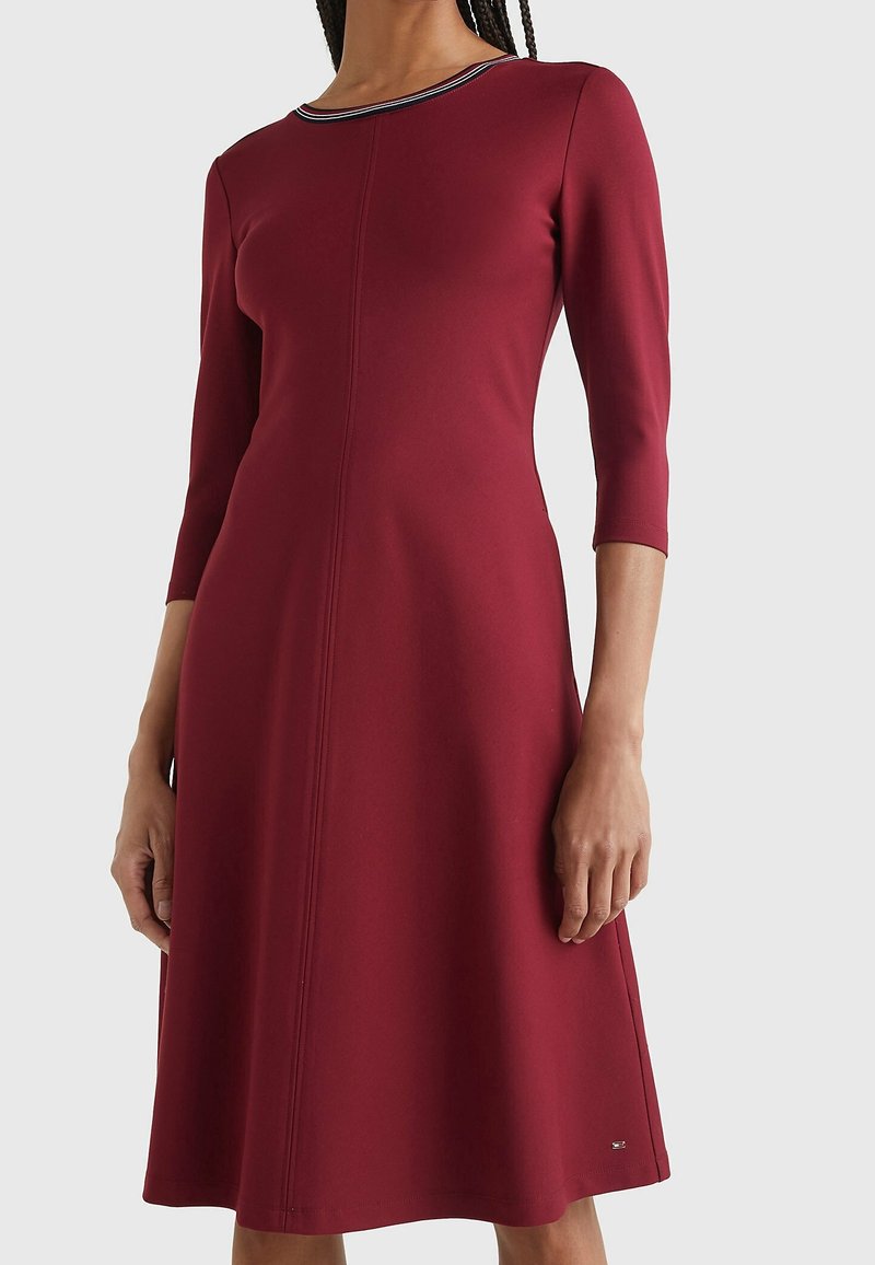 Femme portant une robe bordeaux à manches longues et longueur genou, avec une taille cintrée et un détail rayé au niveau du col.