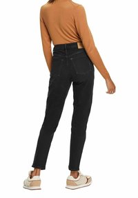 Zwarte jeans met een hoge taille en een taps toelopende pasvorm, voorzien van twee achterzakken en een leren taillebandlabel, gecombineerd met een bruin shirt met lange mouwen.