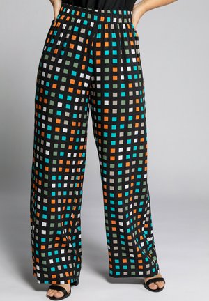 Czarne spodnie o szerokich nogawkach z kolorowym wzorem kwadratów w pomarańczowym, niebieskim, białym i zielonym kolorze, noszone z czarnymi sandałami na otwartym palcu, ręce w kieszeniach.