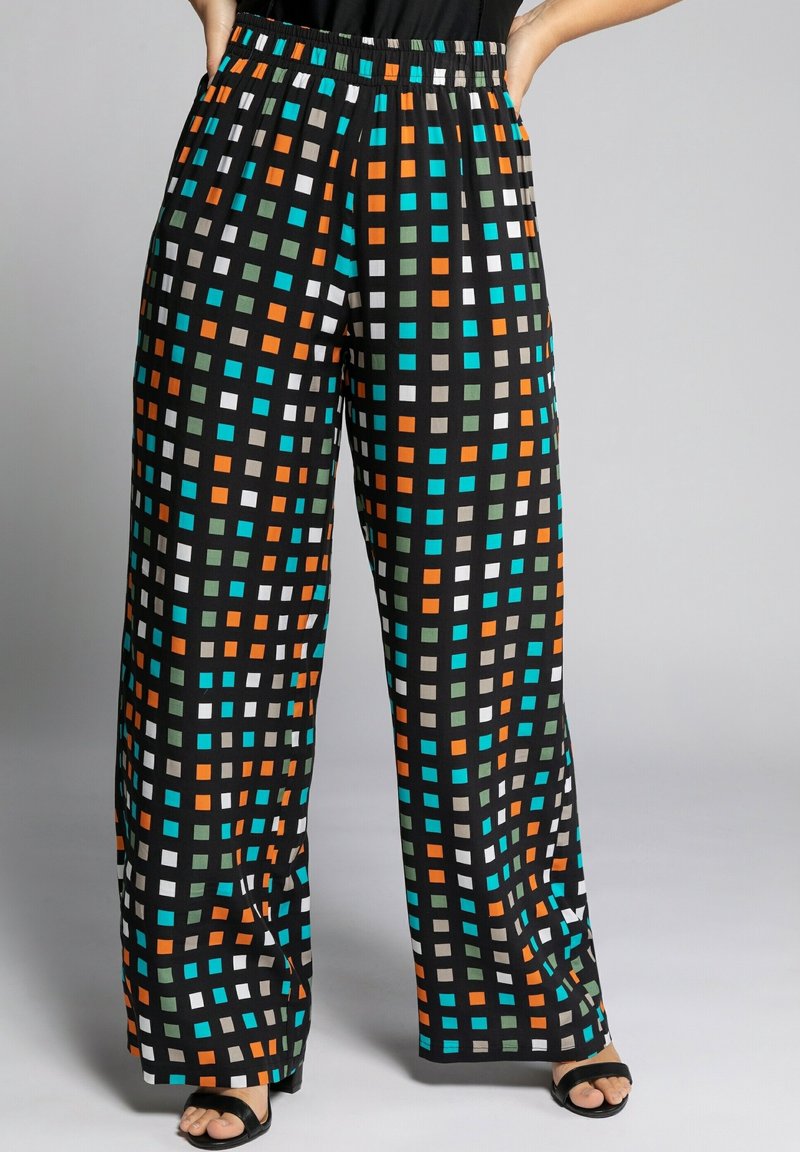 Pantalon noir à jambes larges avec motif carré coloré en orange, bleu, blanc et vert, porté avec des sandales noires à bout ouvert, mains dans les poches.