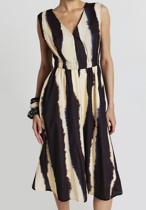Robe midi sans manches avec décolleté en V et taille froncée, ornée de larges rayures tie-dye verticales beiges et noires, portée avec des bracelets imposants.
