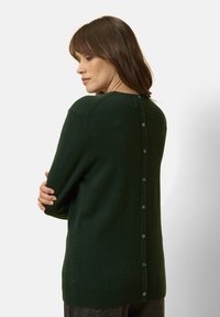 Zelený svetr s jemnou texturou, kulatým výstřihem a knoflíkovým detailem na zádech. Dlouhé rukávy a uvolněný střih dotvářejí design.