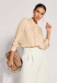 Beige plissiertes Blusenshirt mit Stehkragen, Knopfleiste und geknöpften Ärmeln, kombiniert mit hochtaillierten hellen Hosen und einer taupefarbenen Handtasche.
