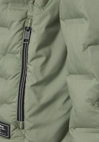 Sekcja oliwkowej kurtki puchowej o pikowanej teksturze, z czarnym zamkiem błyskawicznym i zielonym uchwytem, oraz z etykietą materiałową.