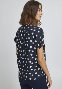 Granatowa bluzka z krótkim rękawem w duże białe kropki, z gładkiego materiału, o luźnym kroju i zaokrąglonym dekolcie. Widoczna subtelna tekstura.
