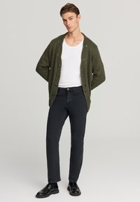 Giovane uomo in piedi con le mani dietro la schiena, indossa una canotta bianca, un cardigan oversized verde oliva, jeans neri e mocassini neri.