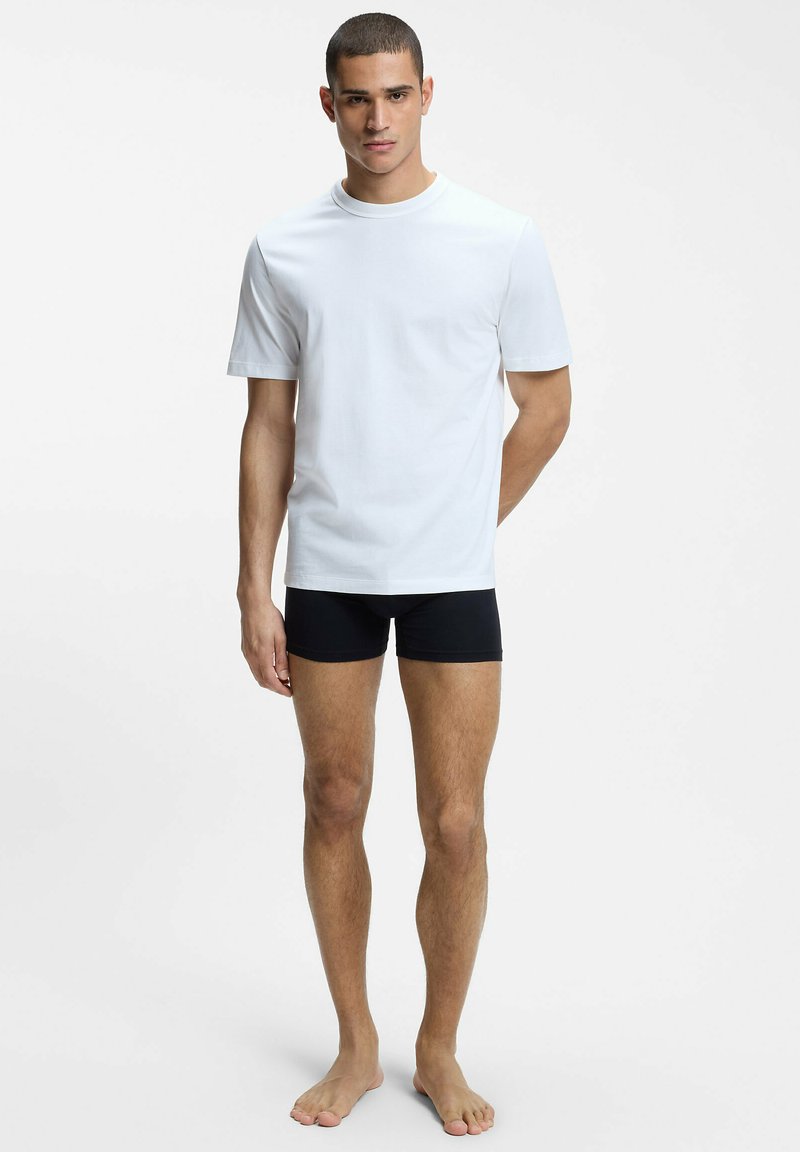 Jeune homme debout pieds nus, portant un t-shirt blanc uni à manches courtes et un boxer noir sur un fond blanc.