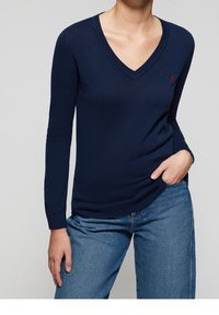 Pull col en V bleu marine en maille légère, avec manches longues et poignets côtelés. Petit logo rouge sur la poitrine gauche.