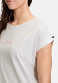 Hvid bomulds-T-shirt med rund halsudskæring og optrukne ærmer med dekorative gyldne hardware-detajler. Subtil flerfarvet print på forsiden.