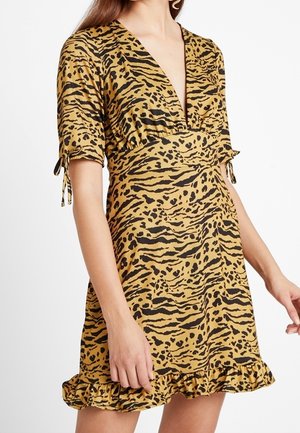 Kobieta ubrana w krótką sukienkę mini o krótkich rękawach, dekolcie w serek, w kolorze musztardowej żółci z czarnym nadrukiem tygrysa i lamparta oraz falbaną na dole, stojąca na białym tle.