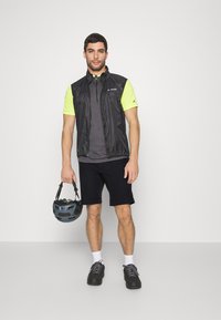 Conjunto de ciclismo para hombre que incluye un chaleco negro, una camiseta de manga corta en amarillo neón, pantalones cortos oscuros y zapatos negros resistentes. Sostiene un casco gris.