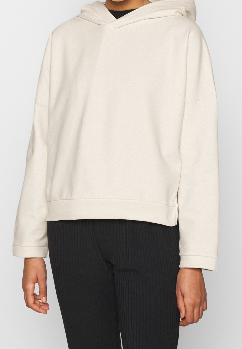 Sudadera con capucha en beige claro de material suave, con bolsillo tipo canguro clásico, ajuste relajado y dobladillo acanalado, combinada con pantalones negros acanalados.