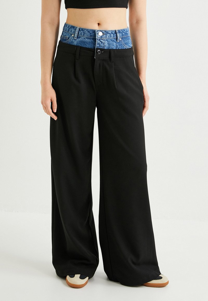 Pantaloni neri a gamba larga con vita in denim, dotati di chiusura a bottone e tasche. Texture liscia, colori contrastanti sulla vita.