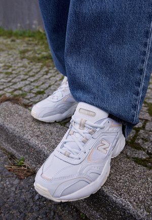 Zapatillas blancas New Balance usadas con vaqueros azules, de pie en el bordillo de una acera de hormigón con pavimento de adoquines cerca.