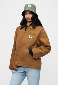 Νεαρή γυναίκα που φοράει καφέ μπουφάν με φερμουάρ και λογότυπο, ανοιχτόχρωμο τζιν και πράσινο καπέλο τύπου bucket με λογότυπο, στέκεται με το ένα χέρι στην τσέπη.