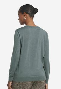 Zielony, długi sweter z długim rękawem, okrągłym dekoltem i ściągaczem. Miękka faktura z gładkim wykończeniem, noszony z wzorzystymi spodniami.