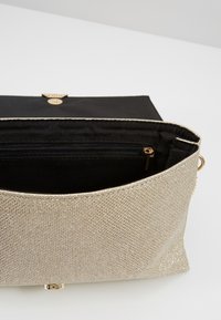 Kopertówka z metalicznej złotej tkaniny z teksturowaną powierzchnią, czarnym wnętrzem i kieszenią na zamek; posiada złote zapięcie na guzik.