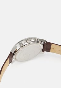 Reloj cronógrafo plateado con caja de metal pulido, correa de piel marrón y dos botones de presión en el lateral. Esfera del reloj texturizada y parte trasera lisa.