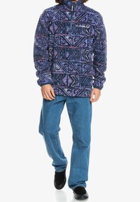 Pullover in pile con mezza cerniera, caratterizzato da un motivo geometrico blu e viola. Abbinato a jeans di denim blu e scarpe nere.