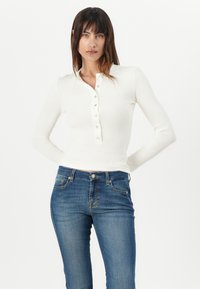 White ribbed long-sleeve top with a buttoned placket, paired with blue denim jeans featuring classic five-pocket design and brass accents.