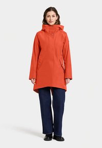 Veste de pluie imperméable orange avec capuche, caractérisée par une coupe droite, des boutons-pression et des poches latérales. Associée à un jean bleu foncé.