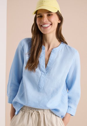 Femme souriante aux longs cheveux bruns porte une casquette jaune, un chemisier bleu clair et un pantalon beige à cordon, sur un fond neutre.